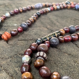 Könnte beinhalten: Eine mehrsträngige Halskette mit einer Vielzahl von Perlen in Braun-, Rot- und Grautönen. Die Perlen bestehen aus Holz, Metall und anderen Materialien. Einige Perlen haben komplizierte Muster und Designs. Die Halskette wird auf einer strukturierten, braunen Oberfläche präsentiert.