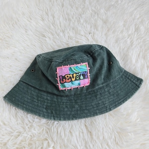 May include: A dark green bucket hat featuring a rectangular patch stitched onto the front. The patch displays a colourful, marbled pattern in shades of pink, blue, and green with the word LEVOTY printed in black letters, surrounded by pink stitching.