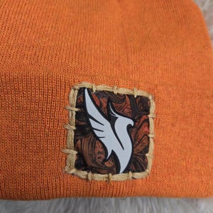 K&ouml;nnte beinhalten: Orangefarbene Strickm&uuml;tze mit einem quadratischen Aufn&auml;her mit einem wei&szlig;en Vogeldesign auf gemustertem Hintergrund. Der Aufn&auml;her ist mit beigem Faden auf die M&uuml;tze gen&auml;ht. Die M&uuml;tze ist aus weichem Strickmaterial.