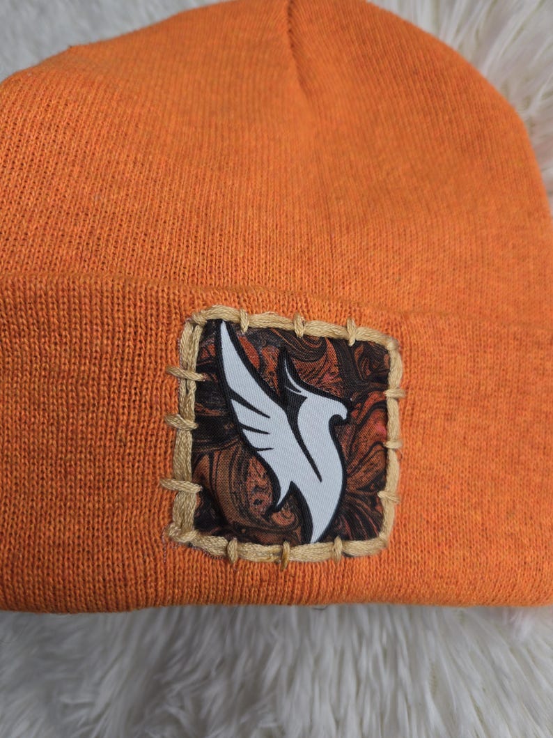 K&ouml;nnte beinhalten: Orangefarbene Strickm&uuml;tze mit einem quadratischen Aufn&auml;her mit einem wei&szlig;en Vogeldesign auf gemustertem Hintergrund. Der Aufn&auml;her ist mit beigem Faden auf die M&uuml;tze gen&auml;ht. Die M&uuml;tze ist aus weichem Strickmaterial.
