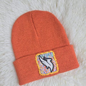 May include: Orange beanie hat with a square patch featuring a white and black wing design on a colorful patterned background. The hat is made of a knitted material.