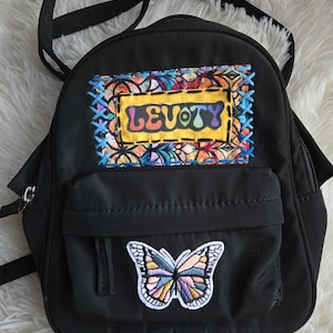 May include: A black mini backpack featuring a rectangular patch with the text LEVOTY in a colorful, psychedelic font, surrounded by a stained-glass pattern and blue cross-stitching. Below, a butterfly patch with pastel-colored wings is centered.