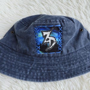 May include: A dark blue denim bucket hat featuring a square patch with a white stylised letter Z graphic over a blue and green aurora borealis background, secured with blue cross-stitch embroidery and two small metal eyelets on the sides.