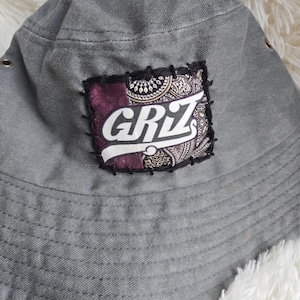 May include: A grey bucket hat featuring a rectangular patch stitched onto the front. The patch displays the text GRIZ in white letters over a purple and brown paisley patterned background, secured with thick black thread stitching around the edges.