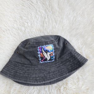May include: A charcoal gray bucket hat featuring a rectangular patch with a stylized white bird graphic against a colorful abstract background. The patch is attached with visible light blue stitching and the hat includes two metal eyelets on the sides.