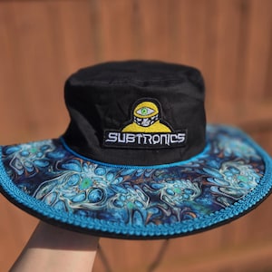 Puede incluir: Sombrero de pescador negro con ala ancha con un patrón azul y turquesa colorido. El sombrero tiene una corona negra y un parche con la palabra "SUBTRONICS" y un gráfico amarillo y verde.