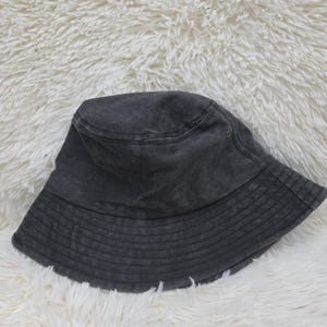 May include: A charcoal gray bucket hat with a washed-out, faded appearance. The hat features a flat, circular crown, a downward-sloping brim with multiple rows of horizontal stitching, and a small metal eyelet on the side for ventilation.