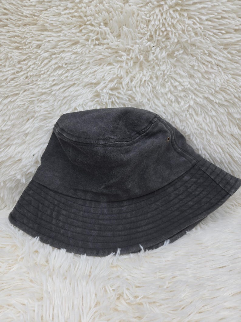 May include: A charcoal gray bucket hat with a washed-out, faded appearance. The hat features a flat, circular crown, a downward-sloping brim with multiple rows of horizontal stitching, and a small metal eyelet on the side for ventilation.