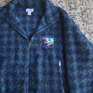 Puede incluir: Una chaqueta de sherpa azul oscuro con textura y cremallera, con cuello alto. La prenda presenta un parche cuadrado en el pecho izquierdo con un diseño gráfico estilizado y costuras moradas. Incluye una cremallera frontal completa y bolsillos.