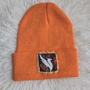 May include: Orange knit beanie with a square patch featuring a white bird design on a brown and tan background. The beanie is folded at the brim and rests on a white, fluffy surface.