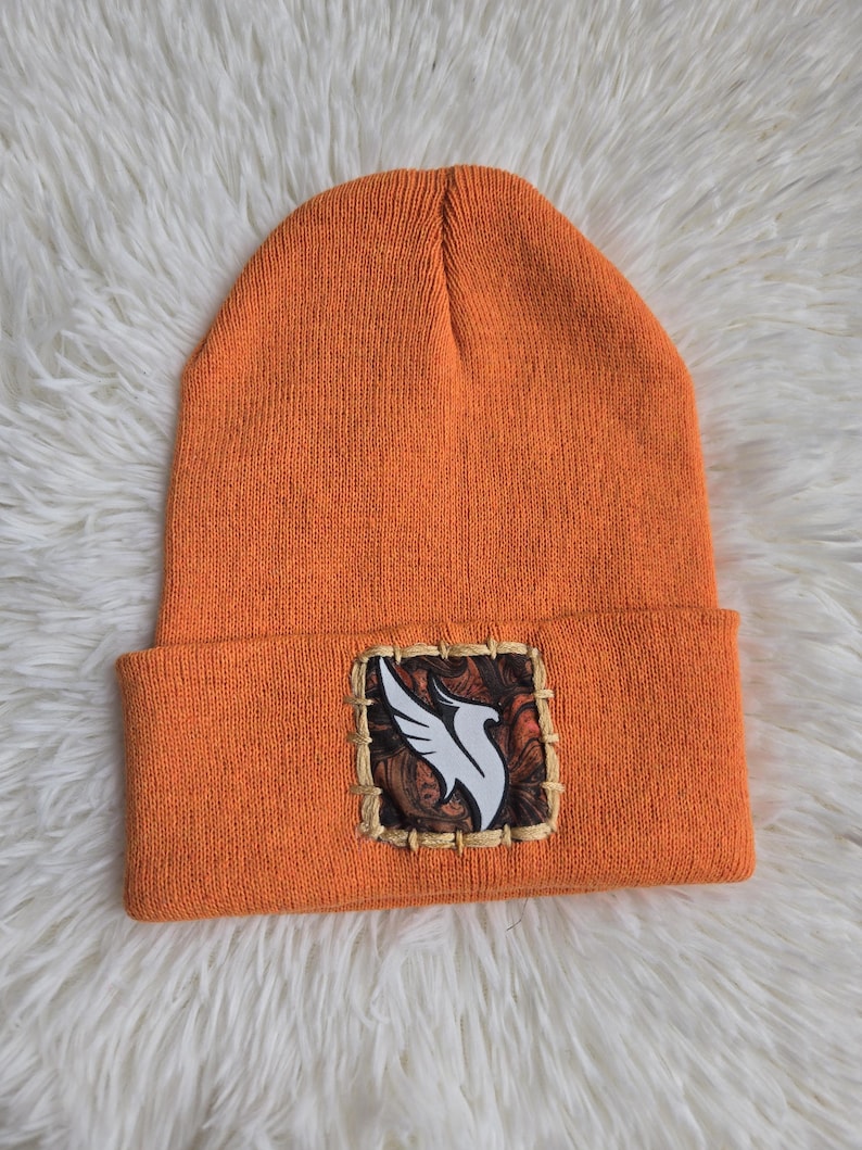 K&ouml;nnte beinhalten: Orangefarbene Strickm&uuml;tze mit einem quadratischen Aufn&auml;her mit einem wei&szlig;en Vogeldesign auf braunem und beigem Hintergrund. Die M&uuml;tze ist am Rand gefaltet und liegt auf einer wei&szlig;en, flauschigen Oberfl&auml;che.