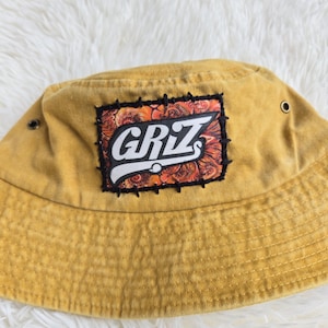 May include: A mustard yellow bucket hat featuring a rectangular patch stitched onto the front. The patch displays the text "GRIZ" in white letters over an orange and black swirling pattern. The hat includes two small metal eyelets on the sides.