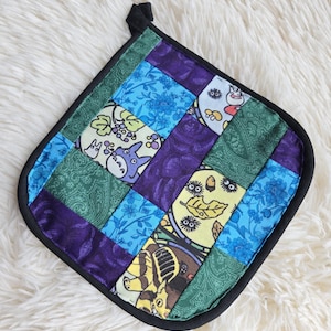 May include: A square quilted potholder featuring a patchwork design of various fabrics. The pattern includes blue floral prints, solid purple fabric, green paisley, and cream-coloured squares with cartoon characters, acorns, and leaves.