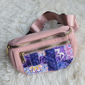 May include: A pink fanny pack with a gold zipper and a patchwork design. The patchwork features various patterns in shades of purple, blue, and white. The adjustable strap is also pink.