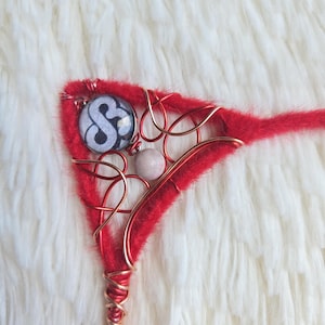 May include: A red hair accessory with copper wire detailing. It includes a circular black and white design, a small pink bead, and wire wrapping. The item is set against a white, fluffy background, creating a striking contrast.