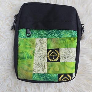 Op de afbeelding: Een zwarte schoudertas met een voorpaneel in een patchworkontwerp van groene en crèmekleurige stoffen. Het patchwork bevat gemarmerde groene patronen, een crèmekleurige stof met kleine bladprints en zwarte vierkanten met een gouden lauwerkrans.