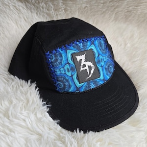 May include: Black baseball cap with a curved brim. The cap features a square patch with a blue and white swirling design and a white graphic. The patch is outlined with blue stitching. A black rope trim borders the patch.