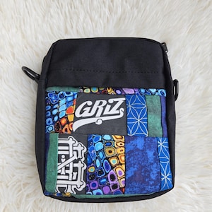 May include: A black crossbody bag featuring a patchwork front panel with various blue, green, and yellow patterned fabrics. The centre displays the white text GRIZ on a dark background, with additional white graphic text on the lower left side.