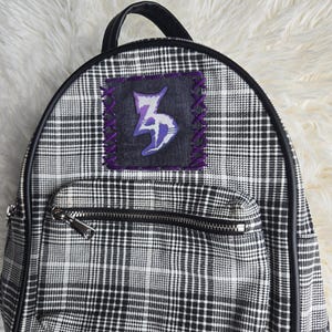 May include: A black and white plaid backpack featuring a black top handle and black trim. The front displays a dark gray rectangular patch with purple cross-stitching and a stylized purple and white graphic symbol. It includes a front zippered pocket.