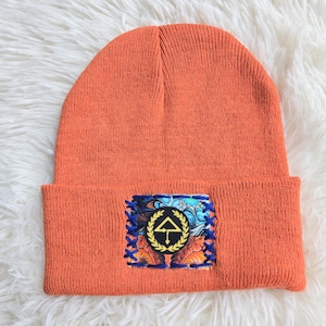 Pode incluir: Um gorro de malha laranja brilhante com um emblema retangular costurado na dobra frontal. O emblema apresenta um círculo preto com uma coroa de louros amarela em torno de uma seta amarela apontada para cima sobre um fundo colorido.