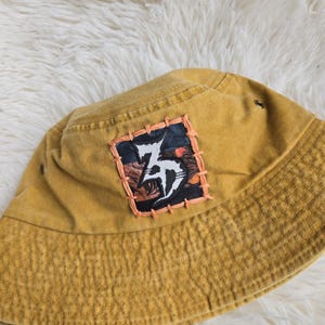 May include: A mustard yellow bucket hat featuring a square patch stitched onto the front. The patch displays a dark, abstract graphic with a prominent white stylised letter design, bordered by thick, light orange hand-stitched thread detailing.