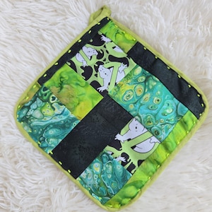 May include: A square quilted potholder featuring a patchwork design of black fabric, green and blue marbled fabric, and a patterned fabric with white ghost figures. The edges are finished with lime green binding and yellow stitching.