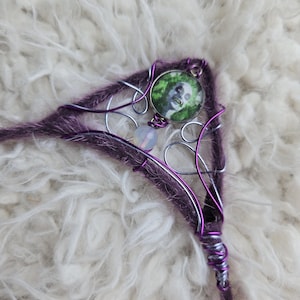 May include: A purple and silver wire wrapped hair stick with a green and white cameo featuring a skull. The cameo is attached to the stick with a silver wire loop.