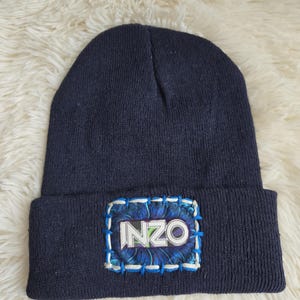 May include: A dark navy blue knit beanie featuring a rectangular patch on the cuff. The patch displays the text INZO in white letters against a blue and black patterned background, secured with visible blue and white decorative stitching.