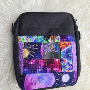 Może przedstawiać: Czarna torba na ramię z przednim panelem wykonanym z kolorowych tkanin w stylu patchwork. Wzory obejmują słońce, różowo-fioletową planetę, siatkę karmazynowych kwadratów oraz geometryczne kształty kwiatowe. Słowo "GOREAM" jest nadrukowane na niebiesko.
