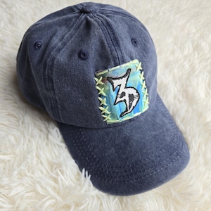 May include: A dark blue denim baseball cap featuring a square patch stitched onto the front panel with light green thread. The patch displays a blue and green abstract painted background with a bold, black and white stylised number three graphic.