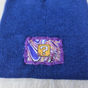 May include: A blue beanie with a sewn-on patch featuring a yellow question mark symbol. The patch has a colorful, abstract design and is outlined with purple stitching. The beanie is made of a soft, knitted material.