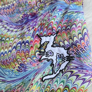 May include: A tote bag with a vibrant, swirling pattern in shades of blue, purple, green, red, and yellow. A white patch with a black outline of a stylized design is sewn on the front. The bag has a blue trim.