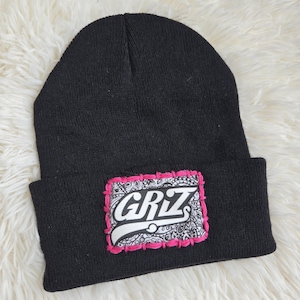Puede incluir: Gorro de punto negro con un parche rectangular. El parche presenta la palabra "GRIZ" en blanco, con un trazo blanco, sobre un diseño de cachemira. El parche tiene un borde rosa.