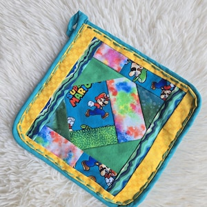 May include: A square fabric potholder featuring a patchwork design with Super Mario character graphics, green fabric panels, and a colorful tie-dye pattern. The border is yellow with a teal trim and a small hanging loop at the top corner.