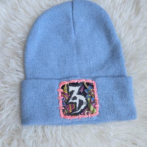 May include: A light blue knit beanie featuring a square fabric patch on the cuff. The patch displays a black and white graphic design with colorful floral patterns, secured to the beanie with thick, bright pink hand-stitched embroidery thread.