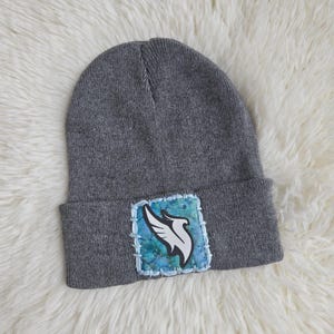 May include: A gray beanie with a folded brim. A square patch with a white bird design and blue and green watercolor background is sewn on the front. The beanie is made of a soft knit material.