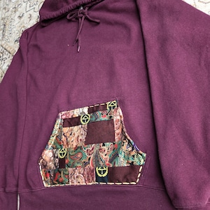 Peut inclure: Un sweat-shirt à capuche bordeaux profond doté d'une poche kangourou frontale composée d'un patchwork de carrés de tissu paisley multicolores et unis. La poche est rehaussée de surpiqûres décoratives jaunes et de petits logos circulaires.