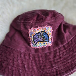 May include: A deep maroon bucket hat featuring a square fabric patch stitched onto the front. The patch displays a colorful, intricate mandala-style pattern with the word "DREAM" embroidered in a stylized, bold black font across the center.