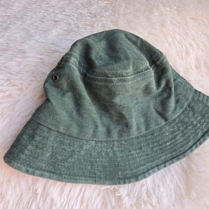 K&ouml;nnte beinhalten: Ein salbeigr&uuml;ner Fischerhut mit breiter Krempe. Der Hut hat eine kreisf&ouml;rmige Krone und ein kleines Taschen Detail. Der Stoff scheint ein weiches, strukturiertes Material zu sein. Der Hut ist ein l&auml;ssiges Accessoire.