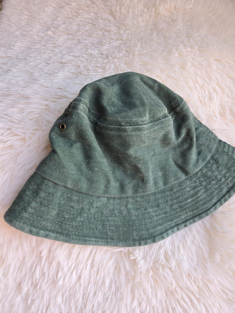 K&ouml;nnte beinhalten: Ein salbeigr&uuml;ner Fischerhut mit breiter Krempe. Der Hut hat eine kreisf&ouml;rmige Krone und ein kleines Taschen Detail. Der Stoff scheint ein weiches, strukturiertes Material zu sein. Der Hut ist ein l&auml;ssiges Accessoire.