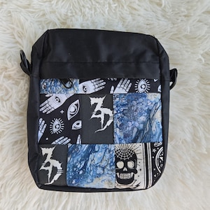 May include: Black crossbody bag with a patchwork design. The patchwork features black and white patterns with eyes, a skull, and abstract designs, along with blue and white marbled fabric. The bag has a black strap and is set against a white, fluffy background.
