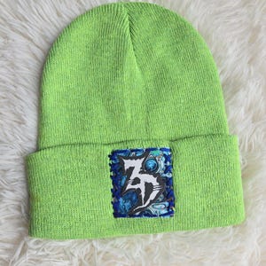 May include: A bright lime green knit beanie with a folded cuff. Centered on the cuff is a rectangular patch featuring a blue and black marbled background with a white, stylized graphic design that resembles a sharp, abstract letter or symbol.