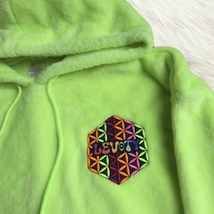 May include: A neon lime green faux fur hoodie featuring a hood with matching drawstrings. On the chest, there is a hexagonal patch with a dark blue background, a multi-coloured geometric flower of life pattern, and the text LEVOTY in purple.
