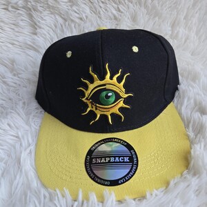 May include: A black snapback cap with a yellow brim and a yellow embroidered sun design featuring a green eye in the center. The brim features a circular sticker with the text SNAPBACK and ORIGINAL SNAPBACK CAP repeated around the edge.