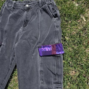 May include: Dark gray cargo pants with white stitching and a purple paisley pocket detail. The pants have a relaxed fit and an elastic waistband. The pocket features a patchwork design with shades of purple and burgundy.