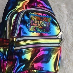 May include: A vibrant, iridescent backpack with a rainbow sheen. The bag features multiple zippered compartments and a patch with the word "LEVOY" in colorful, psychedelic lettering. The bag is set against a fluffy, white background.