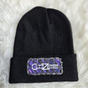 May include: Black beanie hat with a rectangular patch. The patch features the word "CUZZEE" in white, with a purple and gray design. The hat is made of a soft, knitted material.