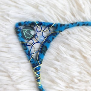 Puede incluir: Un accesorio triangular de oreja de gato con tela de estampado de leopardo azul y negro. El marco está envuelto en alambre metálico azul y dorado, decorado con un pequeño dije circular con un símbolo geométrico y una cuenta translúcida.