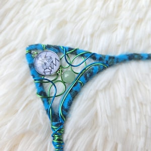 May include: A handmade headpiece with a blue and black patterned fabric base, embellished with green and blue wire. It includes a circular charm with a monochrome design and a small green bead.