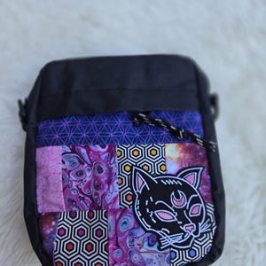 May include: Black crossbody bag with a patchwork design. The bag features a black cat graphic with a crescent moon on the front. The patchwork includes geometric and galaxy-themed patterns in shades of purple, blue, and pink. The bag has a black strap.